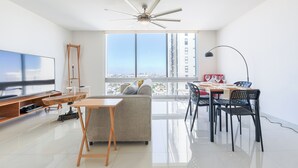 Ein modernes Wohnzimmer mit großem Fernseher, Deckenventilator, Essbereich mit Stühlen und Tisch sowie einem großen Fenster mit Blick auf die Stadt.