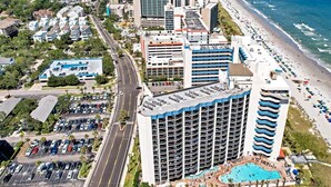 A high-rise hotel with a rooftop pool, adjacent to a beach with umbrellas and people. - Oceanview 1BR Condo • Kid-Friendly Resort • Pools + Arcade (Myrtle Beach)