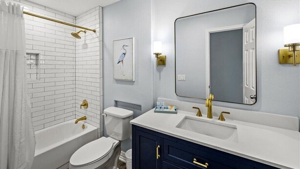 A modern bathroom with white subway tiles, a white toilet, a bathtub with a gold showerhead, a vanity with a white sink and gold fixtures, and a large mirror with a gold frame.