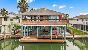 Een tweelaags huis met een balkon, een steiger die in het water reikt, en palmbomen op de achtergrond.