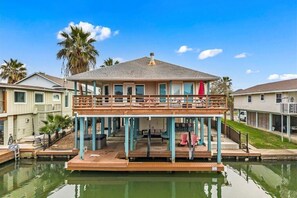 A two-story house with a balcony, a dock extending into a body of water, and palm trees in the background.