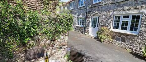 A stone building with a narrow alleyway, a table with a bottle and glasses, and a chair.