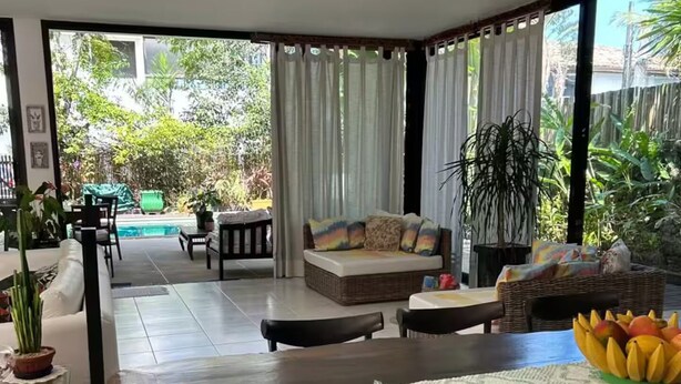 A spacious living area with a wooden dining table, a sofa, and a view of a swimming pool and greenery outside. - Experience Maresias to the fullest: with your feet in the sand, comfort, and exclusivity. (São Sebastião)