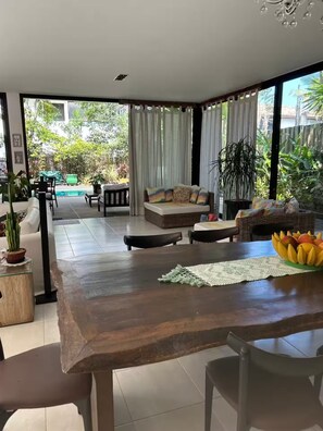 A spacious living area with a wooden dining table, a sofa, and a view of a swimming pool and greenery outside. - Experience Maresia to the fullest: with your feet in the sand, in comfort and exclusivity. (São Sebastião)