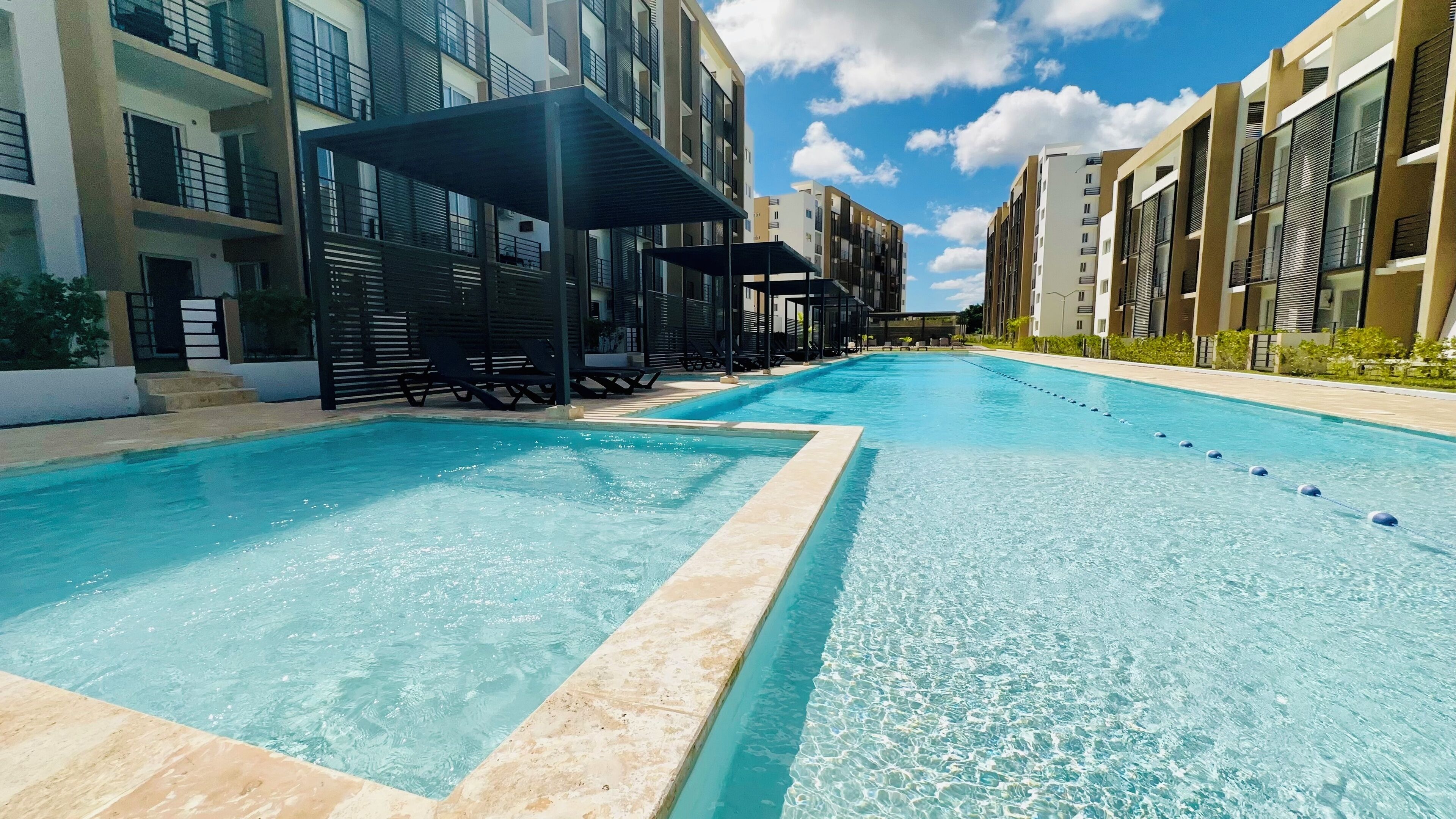 A swimming pool with lounge chairs and umbrellas, surrounded by modern buildings with balconies.