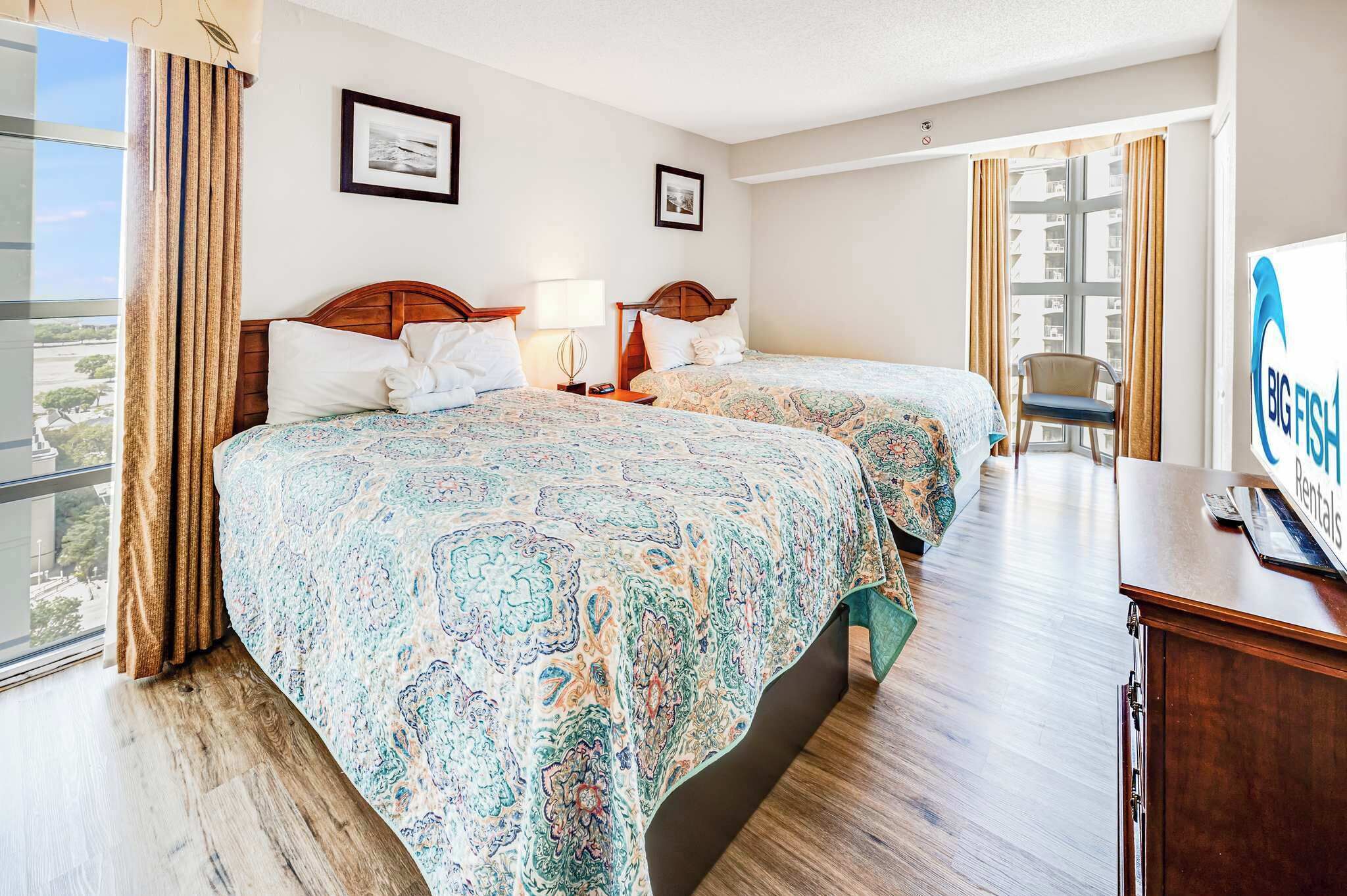 A hotel room with two beds, a desk, a chair, and a large window with curtains.