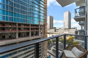 Un balcón con una silla de mimbre, con vistas a una zona de construcción con edificios modernos y una grúa.