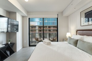 Een moderne hotelkamer met een groot bed, een flatscreen televisie aan de muur, een balkon met uitzicht op een bouwvakpand, en een nachtkastje met een lamp en opgevouwen handdoeken.
