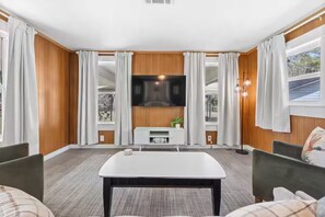 Uma sala de estar moderna com uma televisão de tela plana na parede, uma mesa de centro branca e duas cadeiras.