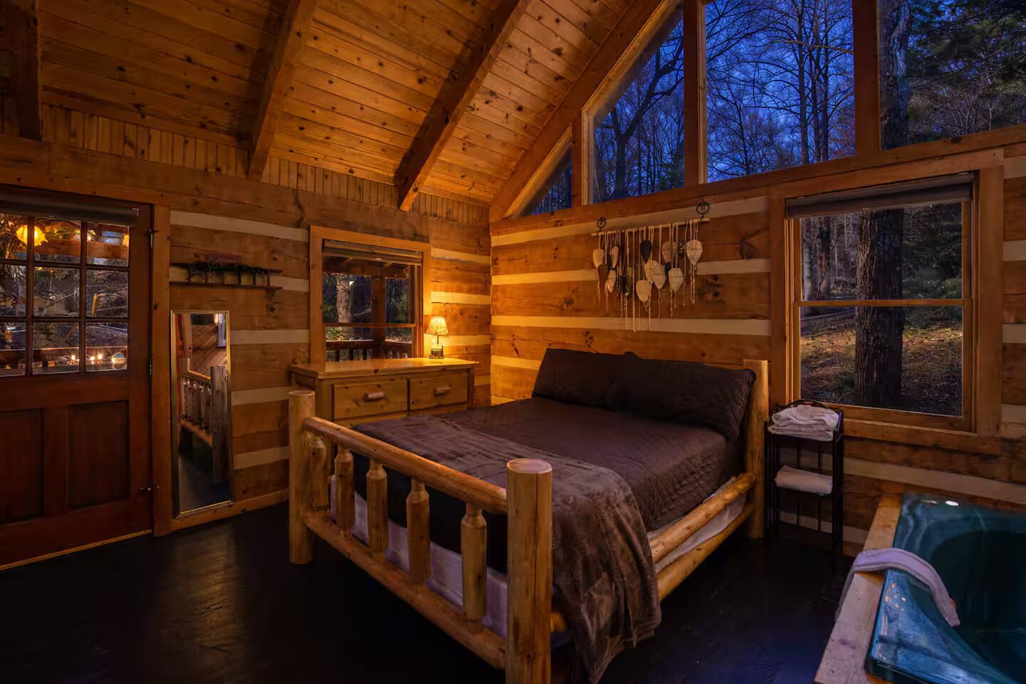 A log cabin bedroom with a wooden bed, a nightstand, a dresser, and a window with a view of trees.