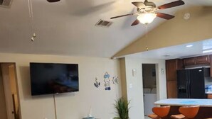 Una sala de estar con televisor, ventilador de techo y área de cocina con taburetes de bar.