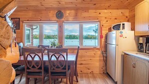 A wooden cabin interior with a dining table, chairs, a refrigerator, and a view of a lake through large windows. - Cozy Paradise - Four Bedroom Cottage, Sleeps 8 (Ryerson)