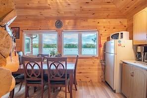 A wooden cabin interior with a dining table, chairs, a refrigerator, and a view of a lake through large windows.