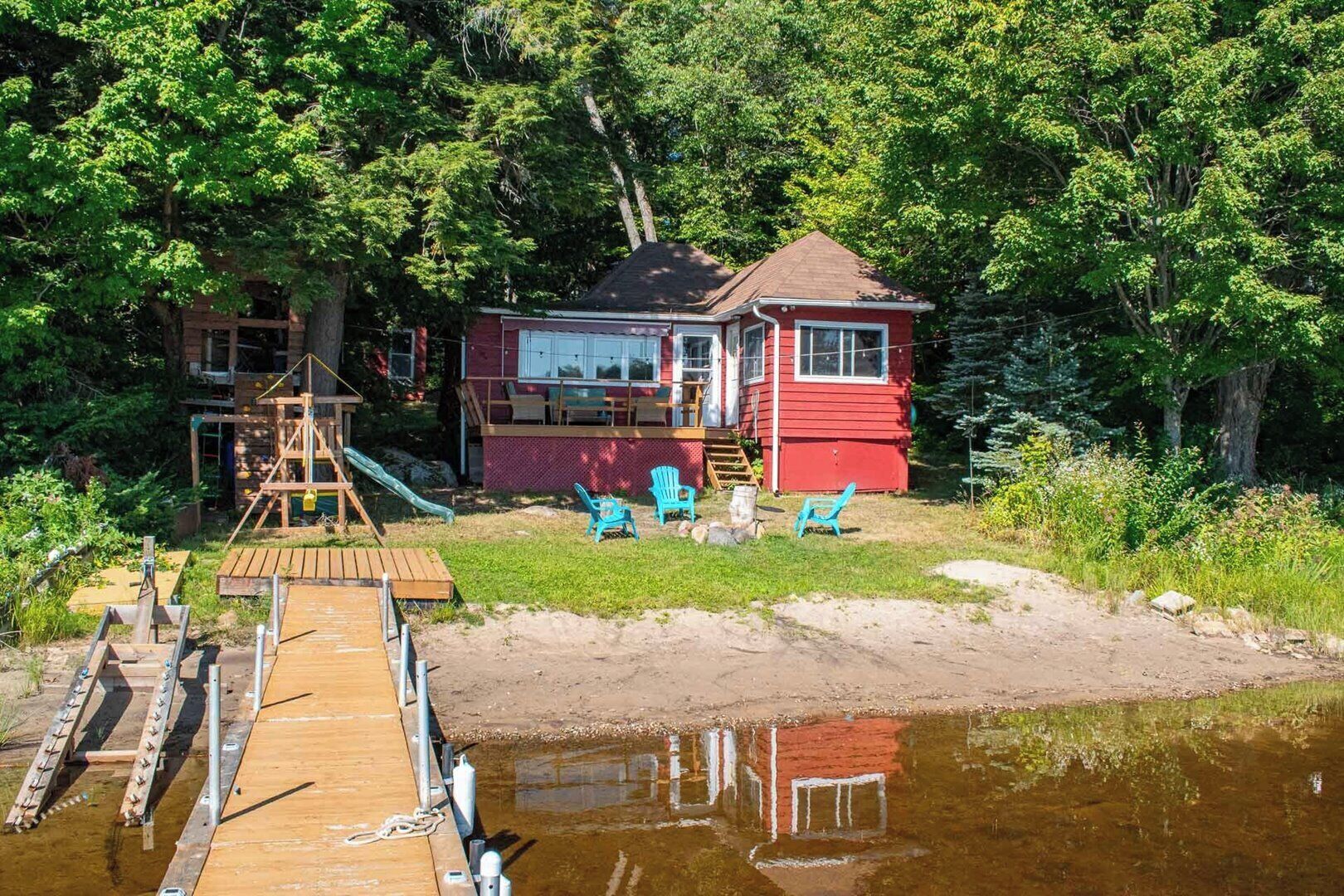 A red cabin with a dock extending into a calm body of water, surrounded by lush greenery.
