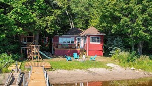 A red cabin with a dock extending into a calm body of water, surrounded by lush greenery. - Cozy Paradise - Four Bedroom Cottage, Sleeps 8 (Ryerson)