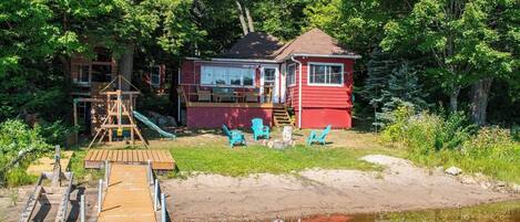 A red cabin with a dock extending into a calm body of water, surrounded by lush greenery.