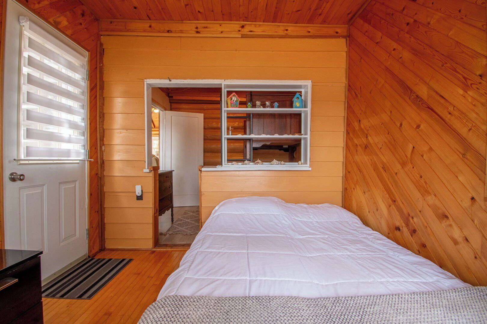 A wooden cabin bedroom with a bed, a dresser, a window with shutters, and a shelf with decorative items.
