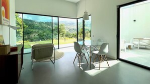 Une pièce moderne avec une table et des chaises en verre, un luminaire suspendu et une vue sur un paysage verdoyant grâce à de grandes fenêtres.