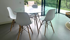 Een moderne eetruimte met een glazen tafel en witte stoelen, grote ramen met uitzicht op een weelderige tuin.