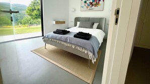 Een moderne slaapkamer met een bed, uitzicht op groen en een kleurrijk abstract schilderij aan de muur.