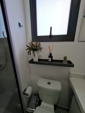 Ein Badezimmer mit Toilette, einer kleinen Ablage mit Pflanze und Flasche sowie einem Fenster über der Toilette.
