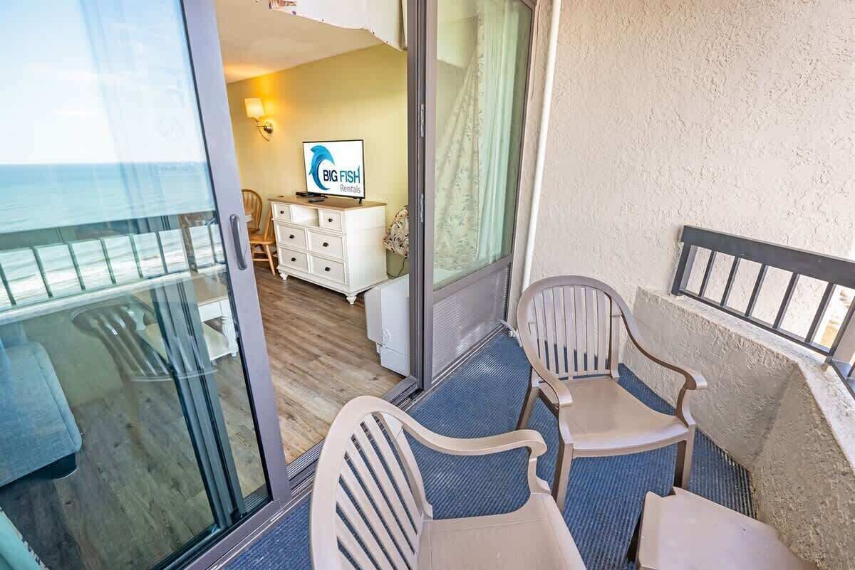 Un balcón con puerta de cristal que da a una habitación con cama, escritorio y televisor mostrando 'BIG FISH Rentals'.
