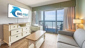 A room with a television displaying 'BIG FISH Rentals,' a sofa, a table, a dresser, and a balcony with a view. - 1BR Compass Cove Oceanfront, Heated Pools, Updated (Myrtle Beach)