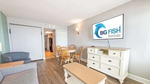 A living room with a sofa, a dining table with chairs, a bench, a white dresser, and a wall-mounted TV displaying 'BIG FISH Rentals'. - 1BR Compass Cove Oceanfront, Heated Pools, Updated (Myrtle Beach)