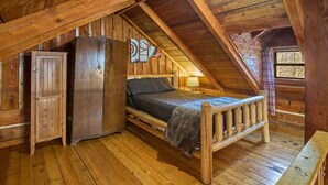 Une chambre mansardée au style rustique, avec un plancher en bois, un lit, un réfrigérateur et une fenêtre donnant sur les arbres.