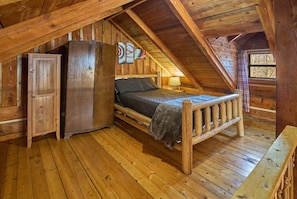 Ein rustikales Schlafzimmer im Dachgeschoss mit Holzböden, einem Bett, einem Kühlschrank und einem Fenster mit Blick auf Bäume.