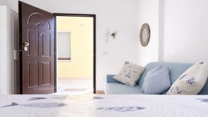 Ein Zimmer mit geschlossener Tür, einem hellblauen Sofa mit gemusterten Kissen, einem Wandspiegel und einem Fenster mit heruntergelassenen Rollläden.