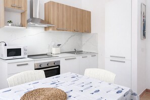 Cocina compacta con armarios blancos, microondas, estufa y pileta. Hay una mesa de comedor con mantel estampado con motivos de peces y dos sillas.