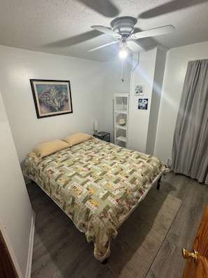 Ein Schlafzimmer mit einem Bett, einem Deckenventilator, einem Nachttisch, einer Lampe und einem gerahmten Bild an der Wand.