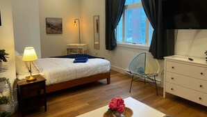 Hotellihuone, jossa on sänky, yöpöytä lampun kanssa, työpöytä tuolilla, lipasto, televisio ja ikkuna, jossa on verhot.
