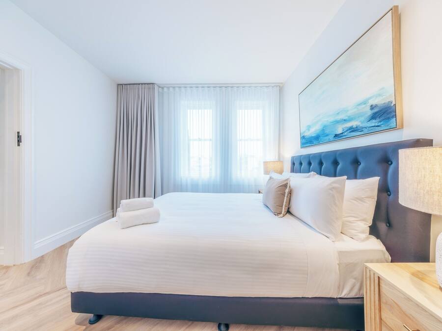 A modern hotel room with a large bed, a headboard with a tufted design, a side table with a lamp, and a painting above the bed.