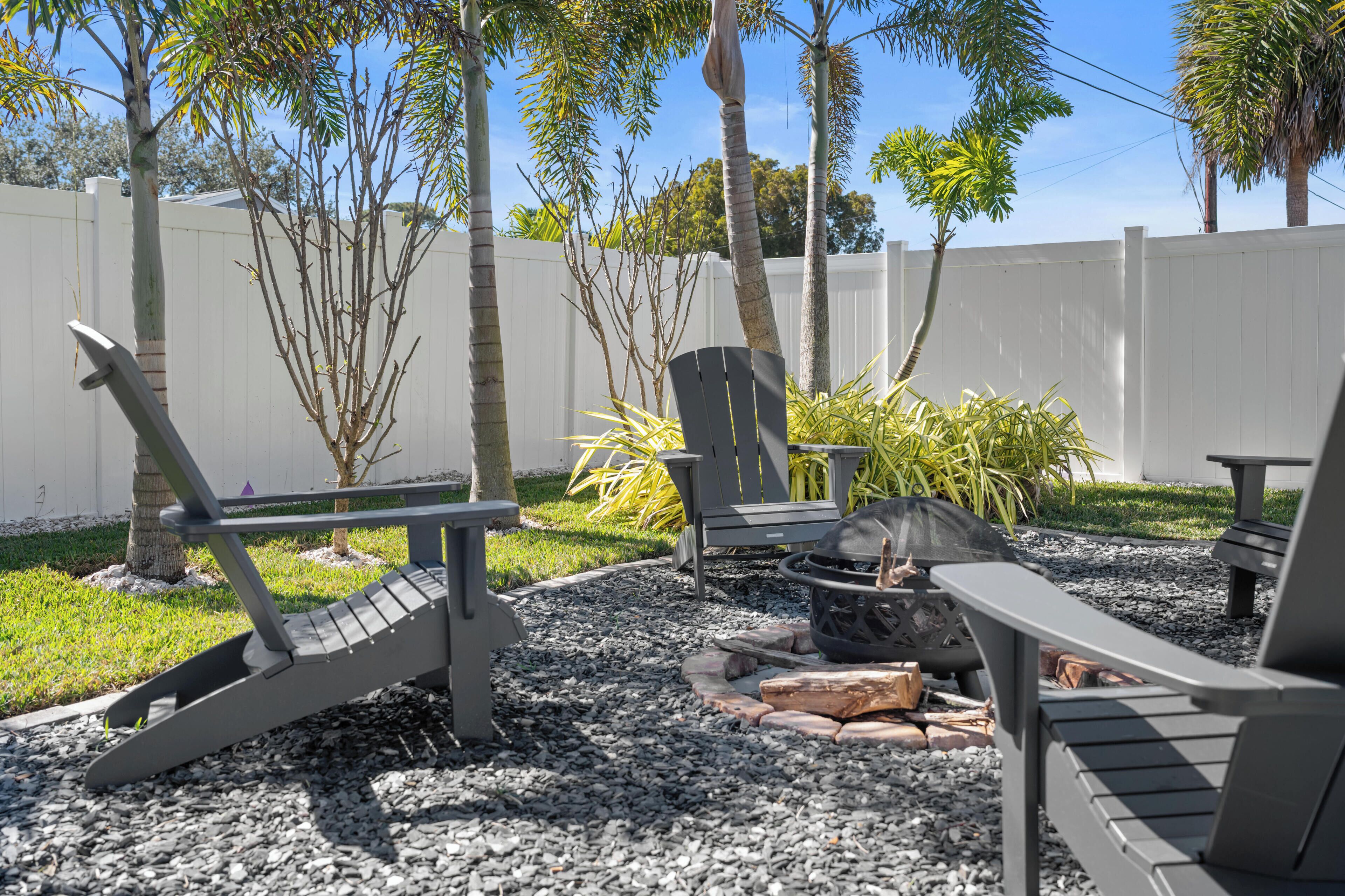 An outdoor seating area with Adirondack chairs, a fire pit, and a white fence in the background.