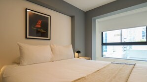 Hotellihuone, jossa on suuri sänky, seinällä kehystetty taulu, yöpöytä lampun kanssa ja ikkuna, josta on näkymä rakennuksiin.