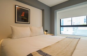Ein Hotelzimmer mit einem großen Bett, einem gerahmten Bild an der Wand, einem Nachttisch mit Lampe und einem Fenster mit Blick auf Gebäude.