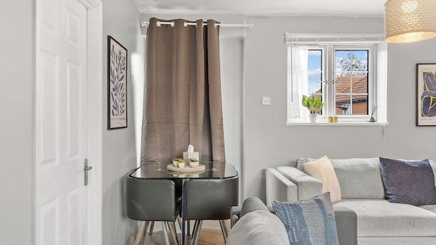 Uma sala de estar moderna com um sofá cinzento, uma mesa de jantar de vidro com duas cadeiras, uma janela com vista para uma casa e uma cortina.