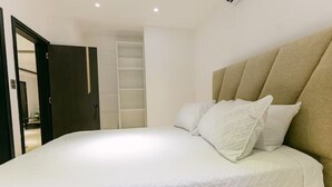 A neatly made bed with white bedding and pillows, a beige headboard, and a built-in shelving unit in the background. - Penthouse con dos camas grande A-301 (Punta Cana)