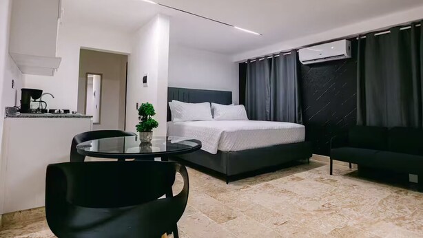 Une chambre d’hôtel moderne comprenant un lit, un canapé noir, une table en verre avec une plante, et un coin cuisine avec une cuisinière.