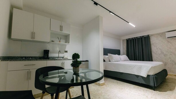 A modern hotel room with a bed, a small kitchenette, a round glass table, and a potted plant. - Habitacion Deluxe cama extragrande A-204 (Punta Cana)