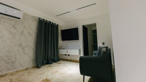 A modern living room with a flat-screen TV mounted on the wall, a black sofa, and a small cabinet below the TV. - Habitacion Deluxe cama extragrande A-204 (Punta Cana)
