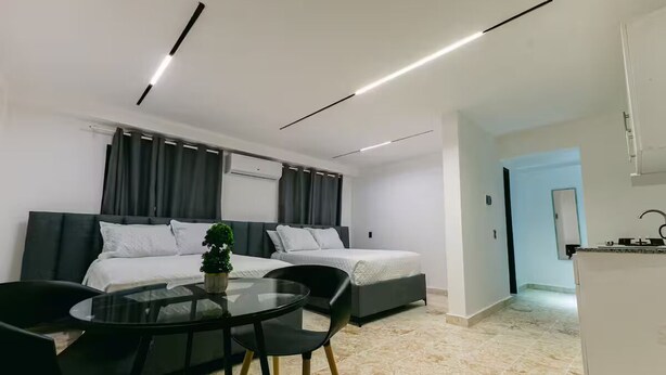Moderni hotellihuone, jossa on kaksi sänkyä, lasinen ruokapöytä, mustat tuolit ja pieni keittiöalue.