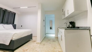 Moderni hotellihuone, jossa on sänky, pieni keittokomero ja kylpyhuone, joka näkyy oven läpi.