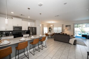 A modern kitchen with a central island, bar stools, a dining area with a table and chairs, a sofa, and a view of the outdoors. - ChampionsGate Resort 5BR Vacation Home | Heated Pool | Minutes to Disney (Kissimmee)