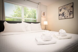 A hotel room with a bed, white linens, a bedside lamp, a painting, and a window with blinds. - ChampionsGate Resort 5BR Vacation Home | Heated Pool | Minutes to Disney (Kissimmee)