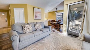 Un salon avec un canapé gris, des coussins à motifs, un coin repas comprenant une table et des chaises, et une porte-fenêtre coulissante donnant sur un balcon extérieur.