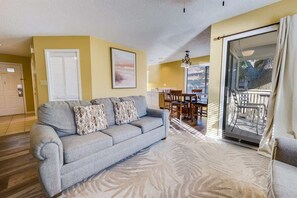A living room with a grey sofa, patterned cushions, a dining area with a table and chairs, and a sliding glass door leading to an outdoor balcony. - New 1BR, Kingston Plantation 11 acre Lake,Location (Myrtle Beach)