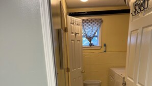 En smal hall med en dörr på vänster sida, ett fönster med mönstrade gardiner, en tvättkorg och en vit toalett.